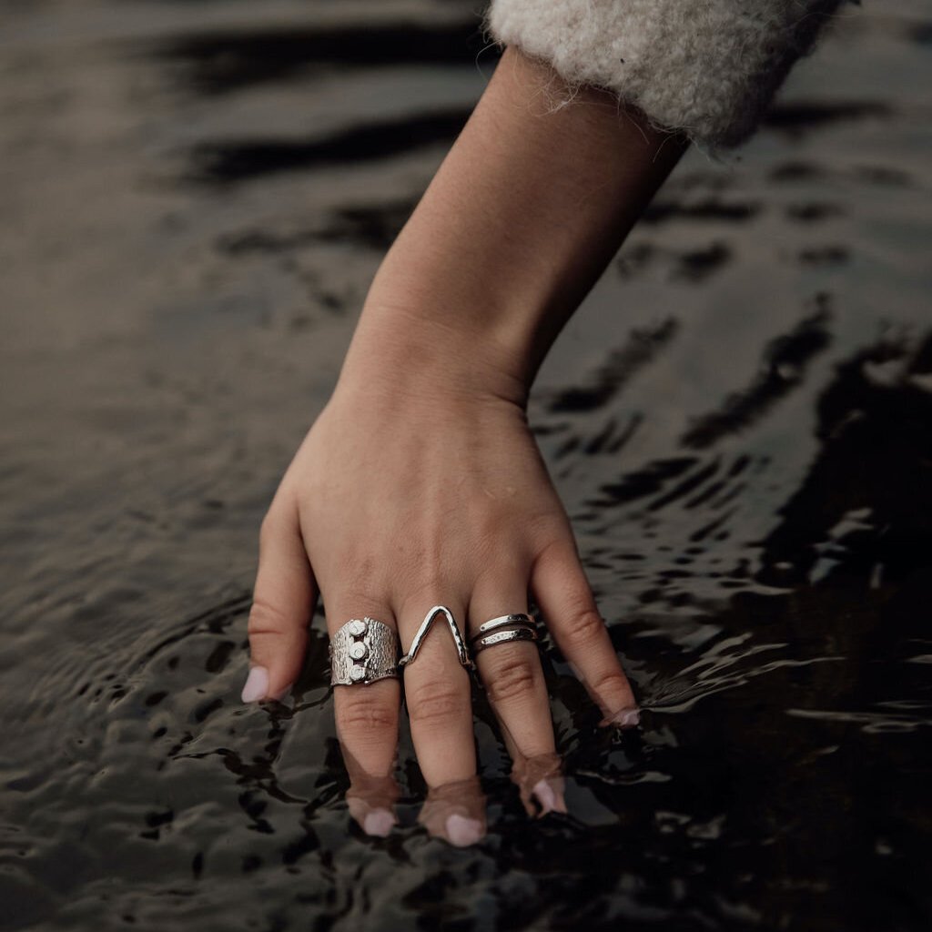 Una mujer sumerge su mano adornada con anillos de plata en un oscuro charco de agua